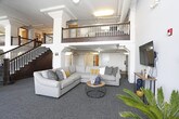 This inviting communal space blends industrial character with modern comfort in a thoughtfully designed open-plan layout. Soaring ceilings with exposed beams and ductwork lend the space an airy, loft-like feel, while a mezzanine balcony with a wood-railed staircase adds vertical drama and architectural interest.

The main floor is organized into distinct yet connected zones. A generously sized lounge area anchors one end of the space, featuring a large sectional sofa in soft grey accented with mustard and neutral throw pillows, arranged around a round wire-and-wood coffee table — a nod to the industrial aesthetic throughout. A wall-mounted flat-screen television completes this relaxed gathering spot.

Adjacent to the lounge, a pair of mid-century-style armchairs sit atop a woven area rug, framed by decorative wall art and a warm floor lamp, creating a quieter reading or conversation nook. A dining area nearby offers a dark wood table and chairs set beside large, multi-pane factory-style windows that flood the space with natural light and offer views of the surrounding neighborhood.

The recreation area features a full-sized pool table bathed in natural light from expansive windows, accompanied by a bench and side table for a comfortable retreat. Throughout, neutral grey carpet, white walls, and structural columns tie the space together into a cohesive, welcoming environment.