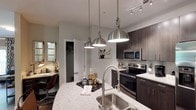 This well-appointed single-floor residence at 3130 Hillandale Rd in Durham, NC, offers a thoughtfully designed layout with modern finishes throughout. The kitchen serves as a central highlight, showcasing dark espresso cabinetry, granite countertops, a large island with an undermount stainless steel sink, and a full suite of stainless steel appliances including a French door refrigerator, electric range, and built-in microwave. Stylish brushed nickel pendant lights hang above the island, adding a contemporary touch. Light wood-style flooring runs through the kitchen and connecting hallways, lending warmth and continuity to the space. The living room is generously sized and bathed in natural light from large windows and a sliding glass door that opens to a private balcony with outdoor views. Crown molding and a ceiling fan add refined detail to this comfortable gathering area. The bedroom is carpeted and serene, with direct balcony access, a ceiling fan, and ample space. The bathrooms include a vanity with granite countertops and dark cabinetry, with one bath featuring a tub-shower combination with tiled surround. A dedicated laundry area houses a full-size side-by-side washer and dryer with overhead wire shelving for added storage convenience.