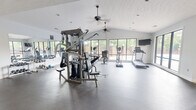 This fitness center is a thoughtfully designed facility comprising a generously sized exercise room and an entry hallway. The main exercise room stands out with its vaulted, textured ceiling accented by dual ceiling fans and recessed lighting, creating a bright and airy atmosphere. Large windows along the exterior wall flood the space with natural light while offering views of surrounding greenery, enhancing the overall workout environment. The rubber-floored exercise area is well-appointed with a comprehensive selection of equipment, including a multi-station cable and weight machine, adjustable weight benches, a full dumbbell rack, treadmills, elliptical trainers, and a stationary bike. A wall-mounted flat-screen television and full-length mirrors further enhance the space, making it functional and motivating. The hallway entry provides a clean transition into the facility, featuring a water fountain, a storage closet, and glass double doors that offer visibility into the main exercise area. The overall ambiance is clean, modern, and practical, with white walls and durable flooring throughout that contribute to a professional fitness environment suited for a variety of workout routines.