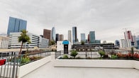 This rooftop terrace delivers a striking urban experience, highlighted by panoramic views of the downtown Los Angeles skyline. The open-air space features a smooth, light-colored concrete deck that provides ample room for a variety of uses and configurations. Sturdy black iron perimeter railings frame the space while preserving unobstructed sightlines of the surrounding cityscape in multiple directions.

Thoughtfully designed raised planters with white stucco exteriors line portions of the perimeter, filled with a curated mix of drought-tolerant agave plants and vibrant flowering red shrubs, adding a touch of greenery against the urban backdrop. A small rooftop structure with a distinctive blue canopy serves as the access point, with an exit door and adjacent lighting providing practical functionality.

The terrace also features additional utility elements, including rooftop mechanical equipment and wall-mounted lighting along the gray stucco access structure. The expansive open deck area is well-suited for events, gatherings, or outdoor activations. Whether taking in the dramatic high-rise skyline to one side or the broader city spread in other directions, this rooftop offers a compelling and versatile outdoor environment in the heart of Los Angeles.