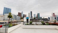 This rooftop terrace delivers a striking urban experience, highlighted by panoramic views of the downtown Los Angeles skyline. The open-air space features a smooth, light-colored concrete deck that provides ample room for a variety of uses and configurations. Sturdy black iron perimeter railings frame the space while preserving unobstructed sightlines of the surrounding cityscape in multiple directions.

Thoughtfully designed raised planters with white stucco exteriors line portions of the perimeter, filled with a curated mix of drought-tolerant agave plants and vibrant flowering red shrubs, adding a touch of greenery against the urban backdrop. A small rooftop structure with a distinctive blue canopy serves as the access point, with an exit door and adjacent lighting providing practical functionality.

The terrace also features additional utility elements, including rooftop mechanical equipment and wall-mounted lighting along the gray stucco access structure. The expansive open deck area is well-suited for events, gatherings, or outdoor activations. Whether taking in the dramatic high-rise skyline to one side or the broader city spread in other directions, this rooftop offers a compelling and versatile outdoor environment in the heart of Los Angeles.