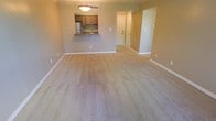 This single-floor unit offers an open and functional layout centered around a spacious living area finished with clean, light-colored carpeting throughout. The warm, neutral-toned walls create a calm and inviting atmosphere, complemented by white baseboards that frame the space neatly.

A defining feature of the unit is the pass-through kitchen, which opens directly into the living area via a raised countertop with a dark stone or laminate surface — ideal as a breakfast bar or casual dining space. The kitchen is equipped with warm wood-toned cabinetry, a stainless steel refrigerator, and a sink positioned beneath the pass-through window, maximizing both functionality and visibility from the main living space. An overhead pendant light fixture illuminates the kitchen bar area, adding a focal point to the open-plan design.

From the living area, a hallway leads further into the unit, suggesting access to additional rooms such as a bedroom or bathroom. A door visible at the end of the hall includes a peephole, indicating it serves as the main entry point. The overall layout is straightforward, efficient, and move-in ready.