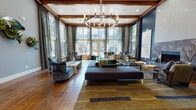 This thoughtfully designed single-floor space blends upscale aesthetics with functional amenities, making it an inviting and versatile destination. The centerpiece is a grand lounge area distinguished by soaring ceilings with exposed wood beam detailing, a coffered white ceiling panel, and rich hardwood flooring that extends throughout. Floor-to-ceiling multi-pane windows flood the room with abundant natural light and offer views of the surrounding greenery, while elegant drapes frame each opening. A striking circular chandelier anchors the space from above, complementing the deep navy blue accent walls that lend a bold, sophisticated character. A tile-surround fireplace anchors one end of the lounge, creating a warm focal point alongside curated seating arrangements and contemporary artwork.

Adjacent to the main lounge, a fully equipped kitchen features dark wood cabinetry, gleaming white countertops, stainless steel appliances including a double oven, microwave, and coffee station, all set against a vibrant teal glass tile backsplash. A peninsula with bar-height seating and pendant lighting makes it ideal for casual gatherings.

A separate lounge nook with arched entryways, a dramatic circular chandelier, and glass-enclosed vanity area adds an intimate, spa-like ambiance. Decorative built-in niches with mirrored backings and floral accents provide elegant finishing touches, completing this polished and welcoming space.