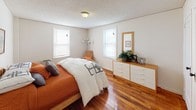 This single-floor residence offers a well-organized layout with warm hardwood flooring flowing throughout the main living areas. The living room is a comfortable, inviting space featuring neutral walls, a textured ceiling, and natural light streaming through windows with blinds. It accommodates a generous seating arrangement, complemented by decorative wall art and accent furniture that add character to the room.

The primary bedroom is notably spacious, featuring two windows that bring in ample natural light, rich hardwood floors, and room for a full dresser and seating area. A second bedroom offers a cozier footprint, also with hardwood floors, a window, and space for a twin bed and desk area, making it well-suited as a guest room or home office.

A compact dining area connects the living space to the kitchen, furnished with a bistro-style table and chairs beneath bold wall art. The kitchen features white cabinetry, dark laminate countertops, a double-basin stainless steel sink, and open shelving for added storage. A bathroom is conveniently accessible from the dining area. Throughout, the consistent hardwood flooring and neutral palette create a cohesive, functional living environment.
