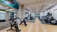 This expansive fitness center offers a comprehensive workout environment thoughtfully divided into distinct functional zones. The space is anchored by prominent natural stone columns that lend a distinctive architectural character, complementing the clean white walls and recessed lighting overhead. A polished hardwood floor runs through the cardio and cycling areas, while patterned carpet tiles define the strength and functional training zones, adding texture and visual variety throughout.

The cardio section is lined with a row of treadmills and elliptical machines positioned along mirrored walls, which enhance the sense of depth and openness. Adjacent to this is a dedicated cycling studio featuring spin bikes arranged on the hardwood floor, with large-scale athletic-themed murals and vibrant graphics adorning the walls, creating an energizing atmosphere.

The strength training area accommodates a full selection of cable machines, free weights, dumbbells, and functional training equipment, including plyo boxes and resistance stations. Vaulted ceilings in portions of the space add to the airy feel, while windows along the perimeter allow natural light to filter in. Large mirrors are strategically placed throughout to support proper form and amplify the overall sense of space. The facility is well-lit, modern, and designed to support a wide range of fitness activities.