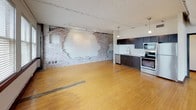 Located at 1510 Main St in the heart of Downtown Dallas, this single-floor space blends industrial character with modern updates. The open layout is anchored by warm hardwood floors that run throughout, complemented by exposed brick walls that add raw, historic texture to the interior. The kitchen is outfitted with dark cabinetry, a white subway tile backsplash, stainless steel appliances including a range, microwave, and dishwasher, and a double-basin sink beneath a pendant light — all set against a structural column that adds to the loft aesthetic. Exposed ceiling infrastructure, including visible ductwork and track lighting, reinforces the industrial design language. The main open area is generously proportioned, offering flexible use potential, and is lined with a row of large windows along one wall that fill the space with natural light. A wall-mounted mirror panel and ceiling fan are also present in this area, suggesting prior use as a studio. The bathroom features hardwood flooring, a dark vanity with a white basin, and a checkered tile tub-shower surround. Throughout the space, the combination of preserved original elements and contemporary finishes creates a distinctive, versatile environment well-suited for a variety of uses.