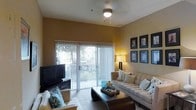 This single-floor living space features a thoughtfully designed layout centered around a comfortable and functional living room. The room is characterized by warm golden-yellow walls that create an inviting ambiance, complemented by neutral-toned flooring and a ceiling fan with a light fixture that provides both comfort and illumination.

A standout feature of the living area is the large sliding glass door fitted with vertical blinds, which floods the room with natural light and offers a view of lush greenery outside, while also providing direct access to an outdoor space. The living room is well-appointed with a plush, light-toned sofa accented with decorative pillows, a dark wood coffee table, and a woven accent chair with tropical-patterned cushions.

A flat-screen television is positioned opposite the seating area, while a floor lamp adds warm ambient lighting in the corner. The wall opposite the sliding doors features a curated arrangement of framed artwork, adding a polished, decorative touch. Overall, the space strikes a balance between comfort and style, making it a welcoming environment for everyday living.