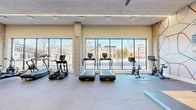 Located at 222 Canal St in New Haven, CT, this well-appointed fitness center offers a spacious, single-floor layout designed to support a full range of workout routines. The space is defined by its striking floor-to-ceiling windows that line the exterior walls, flooding the interior with abundant natural light and providing open views of the surrounding New Haven streetscape.

The cardio zone is prominently positioned along the windowed wall and features multiple treadmills, elliptical trainers, and stationary cycling bikes, allowing users to exercise while enjoying the outdoor scenery. Overhead, sleek linear lighting and ceiling fans maintain a comfortable environment throughout.

The strength training area is comprehensively equipped with a cable machine, adjustable weight benches, a full dumbbell rack, kettlebells, medicine balls, battle ropes, and a rowing machine. Decorative wall murals with geometric and abstract designs add a modern artistic touch to this zone, complementing the contemporary aesthetic of the space.

Exposed concrete columns punctuate the open floor plan, reinforcing the modern industrial design language. The flooring transitions between smooth tile and rubberized surfaces, optimizing each area for its intended use. Accent walls in deep teal add bold color contrast against the neutral warm tones of the surrounding walls, contributing to the vibrant, motivating atmosphere of this well-designed fitness facility.