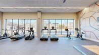 Located at 222 Canal St in New Haven, CT, this well-appointed fitness center offers a spacious, single-floor layout designed to support a full range of workout routines. The space is defined by its striking floor-to-ceiling windows that line the exterior walls, flooding the interior with abundant natural light and providing open views of the surrounding New Haven streetscape.

The cardio zone is prominently positioned along the windowed wall and features multiple treadmills, elliptical trainers, and stationary cycling bikes, allowing users to exercise while enjoying the outdoor scenery. Overhead, sleek linear lighting and ceiling fans maintain a comfortable environment throughout.

The strength training area is comprehensively equipped with a cable machine, adjustable weight benches, a full dumbbell rack, kettlebells, medicine balls, battle ropes, and a rowing machine. Decorative wall murals with geometric and abstract designs add a modern artistic touch to this zone, complementing the contemporary aesthetic of the space.

Exposed concrete columns punctuate the open floor plan, reinforcing the modern industrial design language. The flooring transitions between smooth tile and rubberized surfaces, optimizing each area for its intended use. Accent walls in deep teal add bold color contrast against the neutral warm tones of the surrounding walls, contributing to the vibrant, motivating atmosphere of this well-designed fitness facility.