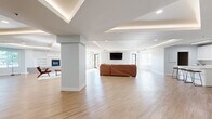 Located at 5560 Library Lane in Colorado Springs, CO, this well-appointed community clubhouse offers a generous single-floor layout designed for both relaxation and recreation. The main gathering area is open and expansive, featuring light-toned hardwood-style flooring, crisp white walls with soft gray accents, and distinctive tray ceilings fitted with elegant indirect LED lighting and recessed fixtures that create a bright, welcoming ambiance throughout.

The lounge area is anchored by a large sectional leather sofa and a wall-mounted flat-screen television, providing a comfortable space for residents to gather. A built-in fireplace with herringbone tile surround and flanking shelving serves as a stylish focal point, while multiple windows allow abundant natural light and views of the surrounding Colorado landscape. A foosball table and additional seating areas cater to a variety of recreational uses.

Structural columns add architectural interest to the open floor plan, subtly defining zones without enclosing the space. A counter-height bar table with stools offers an additional casual seating option, and a kitchenette area with cabinetry is visible toward the rear. A dedicated fitness room rounds out the amenities, equipped with cardio machines for residents' use. The overall design is clean, modern, and highly functional.