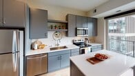 This single-floor studio is designed with an open-concept layout that efficiently combines living, sleeping, and dining areas into one cohesive space. Light hardwood flooring flows throughout, complementing the clean, modern aesthetic.

The kitchen is a standout feature, showcasing flat-front cabinetry in a sophisticated slate-gray finish paired with crisp white countertops. Stainless steel appliances include an electric range, over-the-range microwave, dishwasher, and refrigerator. A peninsula island provides additional prep space and casual seating. An open shelf above the sink adds a decorative touch, ideal for displaying small plants or accents. A sliding glass door adjacent to the kitchen allows natural light to fill the space and provides access to a private balcony.

The living and sleeping area is a generously proportioned open space featuring a ceiling fan and clean white walls. A full bathroom offers a combination tub and shower, a vanity with gray cabinetry, and a sliding barn-style door. In-unit stacked laundry is conveniently tucked within a dedicated closet, adding practical functionality to this well-appointed studio.