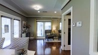 This single-story space showcases a well-appointed interior with rich, dark hardwood flooring that runs throughout, creating a warm and cohesive aesthetic. The living area is a standout feature, highlighted by large windows dressed in vibrant gold curtains that allow generous natural light to fill the room. The neutral gray-toned walls are accented by dark wood crown molding, adding a refined architectural detail that frames the space with character.

The living area is furnished with classic wingback chairs in dark leather, positioned atop a patterned area rug that introduces a pop of color and texture. A table lamp provides warm ambient lighting, complementing the flush-mount ceiling light and recessed lighting overhead. The combination of light sources ensures the space remains bright and inviting at any time of day.

An adjacent room, partially visible through a white-framed doorway, hints at additional living or storage space. A thermostat is conveniently mounted near the entrance, and a console with decorative accessories adds personality to the wall. Overall, the space balances functional design with stylish, contemporary touches.