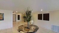 This single-floor space makes a striking first impression with its well-appointed lobby area. At the heart of the entrance stands a large, circular mosaic table crafted from richly patterned stone in earthy tones of green, brown, and cream, serving as a commanding focal point. Atop the table, a tall decorative arrangement of ornamental grasses and botanicals adds a natural, vertical element that draws the eye upward toward the smooth, white ceiling fitted with recessed lighting throughout.

The floors are finished in light-toned ceramic tile, contributing to a clean and open atmosphere. The walls are painted in a warm neutral hue and are adorned with a series of dark-framed decorative panels, lending a sense of symmetry and refined style to the space. A door is visible along one wall, suggesting a practical layout designed for smooth circulation.

The overall ambiance is polished and welcoming, balancing decorative elegance with functional design. The recessed lighting provides even, warm illumination throughout, enhancing the inviting character of the space. Together, the architectural simplicity and carefully selected décor elements create a professional yet approachable environment suitable for a variety of uses.