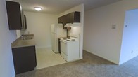 This single-floor residential unit offers a practical and comfortable layout designed for everyday living. The kitchen is efficiently appointed with white appliances, including an electric range and refrigerator, complemented by dark upper cabinetry and an under-cabinet range hood. A dedicated workspace with a sink is positioned nearby, making meal preparation convenient. The kitchen flows naturally into the living area, which is finished with neutral wall-to-wall carpeting that adds warmth throughout the space. The open-concept connection between the kitchen and living room creates a sense of spaciousness and ease of movement. A standout feature of the living area is the sliding glass door that leads to a private balcony, allowing natural light to fill the room and providing an outdoor retreat. An in-unit washer is tucked into a dedicated nook, adding practical convenience without sacrificing living space. Wall-mounted climate control units are present, ensuring year-round comfort. The overall aesthetic is clean and neutral, with light-toned walls and simple finishes that offer a blank canvas for personalization. The layout is straightforward and highly functional, making efficient use of the available space on a single level.