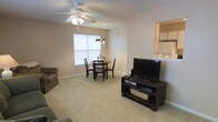 Located at 1601 Wimbledon Dr in Greenville, NC, this single-story home offers a comfortable, open-concept layout that combines the living and dining areas into one cohesive space. The living area features plush wall-to-wall carpeting in a neutral beige tone, contributing to a warm and inviting atmosphere. A ceiling fan with a built-in light fixture anchors the living space, providing both airflow and ambient lighting. The adjoining dining area is positioned near a window that allows natural light to brighten the room, and is accented by a hanging chandelier overhead, adding a modest decorative touch. The walls throughout are finished in a soft, neutral palette that complements the carpeting and keeps the space feeling open and airy. A pass-through or opening visible along one wall suggests a connected kitchen, allowing for easy flow between cooking and living spaces. The overall design is straightforward and functional, with clean lines and a layout well-suited for everyday living. The single-floor plan offers convenient, step-free accessibility throughout the home.