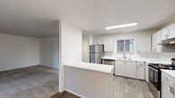 This single-floor residence offers a clean, modern interior with a well-designed open layout. The kitchen is a functional centerpiece, outfitted with white shaker-style cabinetry, white countertops, and stainless steel appliances including a refrigerator, dishwasher, and electric range with an under-cabinet ventilation hood. Wood-look flooring runs throughout the kitchen, which is illuminated by a flush linear ceiling light fixture. A peninsula counter separates the kitchen from the adjacent living and dining areas, which feature neutral carpet flooring.

The living area is bright and open, with a ceiling fan, large windows fitted with blinds, and a sliding glass door that leads to an outdoor deck, providing natural light and exterior views. A hallway connects to the bedrooms, each carpeted and equipped with baseboard heating and windows overlooking the surrounding greenery.

Two bathrooms serve the unit. One features a tub-shower combination with a sliding glass enclosure, while the other includes a standalone shower with a framed glass door. Both bathrooms share a consistent design palette of white cabinetry, white countertops, and wood-look tile flooring, contributing to a cohesive, understated aesthetic throughout the home.