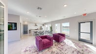 Located at 10742 Freehill Street in Houston, TX, this single-floor space delivers a contemporary, open-concept design that seamlessly integrates a lounge, dining, and kitchen area into one cohesive environment. The interior is finished with light-toned tile flooring throughout, crisp white walls, and recessed lighting that collectively create a bright, clean aesthetic.

The lounge area serves as a central gathering space, featuring a wall-mounted flat-screen television positioned above a sleek linear fireplace set against white tile surround. Tufted magenta armchairs arranged on a patterned area rug add a bold, stylish contrast to the otherwise neutral palette.

The kitchen is well-appointed with granite-topped counters, gray cabinetry, a subway tile backsplash, stainless steel appliances including a range with overhead hood, dishwasher, and refrigerator. A large island with bar-height seating and pendant lighting anchors the kitchen and provides additional workspace and seating.

Multiple large windows and glass doors line the perimeter, flooding the space with abundant natural light and offering direct views of an outdoor pool and surrounding landscape. Gray-framed entry doors lead to exterior access points, reinforcing the functional, multi-use character of this well-designed community space.