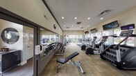 This amenity space in Dania Beach, Florida, consists of a fitness center and lobby area situated within a residential community. The lobby offers a polished, welcoming atmosphere with wood-look flooring, gray walls accented by contemporary artwork, and stylish wingback chairs arranged for comfortable seating. An arched transom window above the entrance door allows natural light to fill the space, while a view-through window connects the lobby to adjacent areas.

The fitness center is accessed through glass-paned French doors and is generously sized, featuring large floor-to-ceiling windows along multiple walls that flood the space with natural light and provide direct views of the outdoor pool area and surrounding landscaping, including palm trees. The room is finished with patterned carpet tile flooring and recessed lighting overhead. Equipment is well-organized and includes treadmills, elliptical machines, stair climbers, and stationary bikes—each featuring individual digital screens. A dedicated free weights section lines one mirrored wall, accompanied by adjustable benches and a cable machine. Multiple wall-mounted flat-screen televisions are positioned throughout for entertainment during workouts. Stability balls and additional accessories round out the functional amenities of this comprehensive fitness facility.