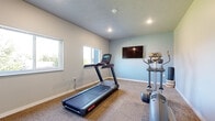 Located at 218 19th St SE in Watertown, South Dakota, this dedicated exercise room offers a practical and well-appointed fitness space. The room features neutral-toned walls in soft gray and blue hues, complemented by carpet tile flooring that provides both durability and comfort underfoot. Recessed lighting illuminates the space alongside natural light streaming in through multiple windows, which also offer views of the surrounding outdoor greenery.

The room is equipped with a treadmill and an elliptical machine, providing a solid foundation for cardiovascular workouts. A wall-mounted flat-screen television adds entertainment value for those looking to stay engaged during exercise sessions. A wall-mounted air conditioning unit ensures climate control for year-round comfort. A yoga mat roll and storage basket are also present, suggesting the space accommodates flexibility and stretching routines as well.

Access to the room is through a keyed entry door featuring a small window panel, offering both security and visibility. The clean, uncluttered layout maximizes the usable floor area, allowing for ease of movement between equipment. The overall design is straightforward and functional, making this exercise room a convenient and comfortable fitness facility.