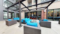 Located at 99 WestEdge Street in Charleston, South Carolina, this sophisticated outdoor courtyard is a standout amenity space designed for both relaxation and social gathering. The centerpiece of the space is a striking black steel pergola structure with a translucent overhead canopy, allowing diffused natural light to filter through while providing shelter from the elements. Beneath the pergola, a sleek fire pit serves as a focal point, surrounded by generously arranged wicker lounge furniture accented with bold turquoise cushions, creating a vibrant and inviting atmosphere. The polished concrete flooring extends throughout the space, lending a modern, refined aesthetic that complements the industrial-inspired framework. Additional seating clusters are thoughtfully positioned throughout the courtyard, encouraging conversation and leisure. Tall palm trees line the perimeter, adding a touch of lush, Southern coastal charm to the setting. The courtyard is enclosed by the multi-story brick and glass building facade, offering a sense of privacy while maintaining an open-air feel. A glass-walled interior lounge is visible and accessible from the courtyard, seamlessly connecting indoor and outdoor living. String lights are strung overhead, enhancing the ambiance during evening gatherings.