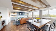 The Sky Lounge at 310 Third St in Farmington, MN is a thoughtfully designed communal gathering space that blends modern comfort with refined, upscale finishes. The open-concept layout seamlessly connects a comfortable living area with a dining space, creating a versatile environment suited for both relaxed socializing and more structured gatherings. Striking exposed wood ceiling beams draw the eye upward and lend a warm, architectural character to the room, complemented by recessed lighting throughout. The living area centers around a rich leather sofa and a fireplace flanked by built-in cabinetry and open shelving in a soft blue-gray finish, providing both storage and display space. A wall-mounted flat-screen TV anchors the lounge area, while a built-in bar area adds an amenity-rich touch. The herringbone-patterned hardwood flooring flows throughout the entire space, adding texture and visual depth. Large windows along multiple walls flood the interior with abundant natural light and frame views of the surrounding Farmington landscape. A sliding glass door opens directly onto an outdoor terrace, extending the usable space and offering elevated open-air views. The dining area accommodates a large dark-wood table with ample seating, making the Sky Lounge an ideal setting for community events and private gatherings alike.