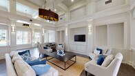 This sophisticated single-floor community space showcases an impressive blend of classical architectural design and modern comfort. Soaring double-height ceilings with detailed coffered and tray moldings immediately command attention, amplifying the sense of grandeur throughout the interior. Crisp white wainscoting, decorative pilasters, and built-in niches line the walls, lending a refined, formal character to the space.

The expansive main lounge area is anchored by a central seating arrangement featuring neutral linen sofas and armchairs accented with navy and cobalt blue pillows, set atop a woven gold area rug over rich dark-toned tile flooring. A large flat-screen television is mounted within a paneled built-in, flanked by wall sconces that provide warm, layered lighting throughout.

A welcoming entry foyer with floor-to-ceiling windows and glass double doors floods the space with natural light, while artwork and decorative accents add curated personality. Adjacent to the lounge, a casual café-style seating area with bistro tables and stools sits beside oversized windows that frame panoramic views of a resort-style swimming pool and the surrounding community. A glimpse into a neighboring fitness room further highlights the multifunctional nature of this well-appointed amenity space.