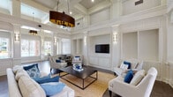 This sophisticated single-floor community space showcases an impressive blend of classical architectural design and modern comfort. Soaring double-height ceilings with detailed coffered and tray moldings immediately command attention, amplifying the sense of grandeur throughout the interior. Crisp white wainscoting, decorative pilasters, and built-in niches line the walls, lending a refined, formal character to the space.

The expansive main lounge area is anchored by a central seating arrangement featuring neutral linen sofas and armchairs accented with navy and cobalt blue pillows, set atop a woven gold area rug over rich dark-toned tile flooring. A large flat-screen television is mounted within a paneled built-in, flanked by wall sconces that provide warm, layered lighting throughout.

A welcoming entry foyer with floor-to-ceiling windows and glass double doors floods the space with natural light, while artwork and decorative accents add curated personality. Adjacent to the lounge, a casual café-style seating area with bistro tables and stools sits beside oversized windows that frame panoramic views of a resort-style swimming pool and the surrounding community. A glimpse into a neighboring fitness room further highlights the multifunctional nature of this well-appointed amenity space.