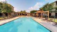 This outdoor amenity space centers around a generously sized resort-style swimming pool with clear, blue water that immediately sets a relaxing and inviting tone. The pool deck is finished with a light-toned surface and lined with multiple lounge chairs, offering ample space for sunbathing and leisure. A handsome wooden pergola anchors one side of the pool area, providing shaded seating and an attractive architectural focal point that adds warmth and character to the setting. Mature palm trees and well-maintained greenery frame the space, contributing to a lush, tropical atmosphere. Additional seating and dining areas are visible near the surrounding building, which features a single-story layout with a neutral, earth-toned exterior and a welcoming facade. The grounds are thoughtfully landscaped, with trees and shrubs creating a sense of privacy and seclusion. The open sky above and the abundance of natural light enhance the overall ambiance, making this outdoor space feel bright, airy, and ideal for relaxation and outdoor gatherings. The combination of the pool, pergola, and verdant surroundings creates a cohesive and appealing outdoor retreat.