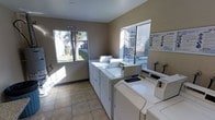 This single-floor shared laundry facility is designed for practical, everyday use and offers a clean, functional environment for residents. The room is outfitted with multiple full-sized top-loading washers and dryers arranged along the walls, providing ample capacity for laundry needs. The light tan tile flooring is durable and easy to maintain, contributing to the room's tidy, utilitarian character.

Two windows allow generous amounts of natural light to fill the space, with views of the surrounding outdoor area including mature trees and greenery, giving the room an open, airy feel despite its compact size. The neutral-toned walls keep the atmosphere clean and straightforward.

Informational signage is posted on the walls, including usage instructions and facility rules, ensuring users have clear guidance while utilizing the equipment. A no-smoking sign is also prominently displayed, reinforcing a clean and safe environment. The overall ambiance is bright, orderly, and purpose-driven, making it a convenient and accessible amenity for shared use.
