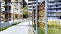 This outdoor courtyard is a well-appointed communal space that seamlessly blends modern architectural design with natural elements. The area is defined by a series of steel-framed pergolas accented with warm-toned hardwood slat panels, which serve both as decorative privacy screens and structural design features, creating a sense of enclosure while maintaining an open, airy feel.

The hardscaped pathways, finished in large-format light grey pavers, wind through the space and are complemented by decorative gravel borders and thoughtfully placed ornamental grasses and native plantings. Raised planter beds and curated greenery add layers of texture and color throughout, contributing to a serene, park-like atmosphere.

The courtyard is enclosed by the surrounding multi-story building, whose façade combines warm brick tones with large windows and private balconies, providing an architectural backdrop that enhances the sense of place. Natural light floods the space, highlighting the interplay between the steel, wood, and landscaped elements.

Overall, the courtyard offers a refined outdoor retreat that balances urban sophistication with natural warmth, making it an inviting space for relaxation and casual gathering.