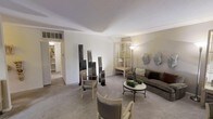 This single-floor space presents a thoughtfully curated interior defined by a sophisticated, neutral color palette of soft whites, warm beiges, and muted grays. The main living area serves as the centerpiece, showcasing a harmonious blend of modern and classic design sensibilities. The room features plush carpeted flooring and smooth white ceilings, lending an overall sense of calm and refinement.

A velvet upholstered sofa anchors the seating arrangement, complemented by a pair of cream-toned armchairs with clean lines. A round marble-top coffee table with a dark wrought-iron base adds an elegant contrast to the soft furnishings. Decorative greenery on the table introduces a subtle organic touch.

Artistic floor-standing sculptural panels in dark metal create a striking visual focal point, adding depth and creative character to the space. A glass display cabinet with warm interior lighting provides both functional storage and a decorative accent. Wall-mounted sculptural pieces in muted tones further enhance the gallery-like ambiance.

A hallway visible in the background leads to additional areas of the space, featuring built-in shelving. Recessed HVAC venting is neatly integrated into the ceiling, maintaining the clean aesthetic. The overall atmosphere is serene, polished, and artistically expressive.