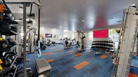 This spacious fitness center is designed to support a comprehensive workout experience, combining a modern aesthetic with a wide array of equipment. The floor is covered in a striking blue carpet accented with bold orange panels, lending the space a vibrant, energetic atmosphere. Soaring ceilings — including a coffered section with pendant lighting — amplify the sense of openness throughout.

The cardio zone features multiple treadmills, ellipticals, a rowing machine, stair climbers, and stationary bikes, all positioned to take advantage of the large floor-to-ceiling windows that offer outdoor views of the surrounding courtyard and pool area. Natural light fills this section of the room, creating an inviting environment for cardio workouts.

The strength training area is equally well-appointed, housing a multi-station cable machine, a functional training rig, weight benches, and an extensive collection of dumbbells and kettlebells neatly organized on wall-mounted racks. Full-length mirrors line one wall, enhancing both the sense of space and practicality for form-checking during workouts.

Wall-mounted flat-screen televisions and a large digital display screen provide entertainment and motivation throughout. Glass partition walls near the entrance connect the fitness center to an adjacent common area, maintaining visual openness while defining the space. Recessed lighting supplements the natural light for a well-lit, welcoming environment at any time of day.
