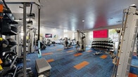 This spacious fitness center is designed to support a comprehensive workout experience, combining a modern aesthetic with a wide array of equipment. The floor is covered in a striking blue carpet accented with bold orange panels, lending the space a vibrant, energetic atmosphere. Soaring ceilings — including a coffered section with pendant lighting — amplify the sense of openness throughout.

The cardio zone features multiple treadmills, ellipticals, a rowing machine, stair climbers, and stationary bikes, all positioned to take advantage of the large floor-to-ceiling windows that offer outdoor views of the surrounding courtyard and pool area. Natural light fills this section of the room, creating an inviting environment for cardio workouts.

The strength training area is equally well-appointed, housing a multi-station cable machine, a functional training rig, weight benches, and an extensive collection of dumbbells and kettlebells neatly organized on wall-mounted racks. Full-length mirrors line one wall, enhancing both the sense of space and practicality for form-checking during workouts.

Wall-mounted flat-screen televisions and a large digital display screen provide entertainment and motivation throughout. Glass partition walls near the entrance connect the fitness center to an adjacent common area, maintaining visual openness while defining the space. Recessed lighting supplements the natural light for a well-lit, welcoming environment at any time of day.