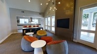 This contemporary single-floor community space is thoughtfully designed to encourage both relaxation and social interaction. The room immediately draws the eye upward with its dramatic vaulted ceilings, anchored by a striking sputnik chandelier with exposed Edison-style bulbs that serves as a bold focal point. The open-plan layout seamlessly connects a casual lounge area with a fully equipped kitchen space in the background.

The lounge area is furnished with a curated collection of rounded, low-profile seating in vibrant orange, yellow, and grey tones, paired with sleek white accent tables — creating an inviting and energetic atmosphere. A large wall-mounted flat-screen display is set against a textured wood-panel accent wall, ideal for gatherings or presentations.

The kitchen area at the rear features a long bar-height dining counter with bold red seating, complemented by dark cabinetry and a statement range hood, making it well-suited for entertaining or communal dining. Tall French doors along the back wall flood the space with natural light and provide direct access to an outdoor area, enhancing the sense of openness. The overall ambiance is modern and lively, with warm tones and thoughtful design details throughout.