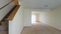 Located at 2332 Asics Dr. in Hilliard, Ohio, this two-story home offers a comfortable and functional layout spread across two levels. The main living area is visible in the image, showcasing a spacious, open floor plan with neutral wall-to-wall carpeting that extends throughout the connected rooms, creating a cohesive and understated aesthetic.

The living space is characterized by clean white walls that lend a bright, airy feel to the interior. A partial wall divides the main area from an adjoining room, which benefits from natural light streaming through a large window dressed with sheer white curtains. A ceiling-mounted light fixture serves this secondary space, providing additional illumination.

A notable architectural feature is the staircase situated near the entry area, accompanied by built-in wooden shelving that adds a warm, functional accent to the otherwise neutral palette. The staircase leads to the second floor, expanding the overall living space of the home.

Visible HVAC vents in the ceiling indicate a central air system throughout. The overall ambiance is clean, neutral, and move-in ready, offering a blank canvas well-suited for personalization.