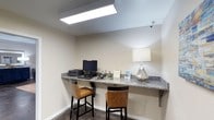 This business center is a focused, efficiently designed workspace that provides essential amenities for work and productivity. The centerpiece of the room is a wraparound granite countertop workstation, offering ample surface area for multiple users simultaneously. Two cushioned bar-height chairs with warm tan upholstery and sleek black frames are positioned at the counter, providing comfortable seating during work sessions. The workstation is equipped with a desktop computer and printer, making it convenient for tasks such as document printing and internet access. A decorative lamp with a geometric metallic base and a neutral fabric shade adds a touch of refined style to the space while contributing supplemental ambient lighting. The room is illuminated by a large overhead fluorescent panel light that ensures bright, even lighting throughout. The walls are finished in a clean, neutral tone, and the flooring features dark tile, lending a polished, professional appearance. A security camera is mounted near the ceiling, ensuring a safe and monitored environment. Overall, the space is purposefully arranged to support productivity in a clean, professional setting.