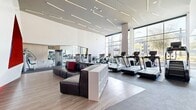 This expansive 3,290-square-foot fitness center at 1912 S Jacaranda Street in Anaheim, California, is a single-floor facility designed with a sleek, modern aesthetic. The space is defined by soaring ceilings, floor-to-ceiling glass walls, and linear LED lighting that creates a bright, energizing atmosphere throughout. Natural light pours in from large windows and a glass garage door, offering outdoor views of palm trees and landscaped surroundings. The cardio zone features a well-equipped row of treadmills, ellipticals, and stationary cycling bikes positioned to maximize natural light and views. A dedicated strength training area houses Freemotion cable machines and selectorized equipment on cushioned carpet flooring, complemented by full-length mirrors. The functional training zone includes a TRX rig, heavy bag, battle ropes, and a full set of KAOS free weights on durable rubber flooring, alongside a bold large-scale athletic mural. A welcoming lounge area with modular seating and a vibrant red and white geometric accent wall with an embedded display screen adds a dynamic social focal point. Warm wood paneling, multi-toned flooring transitions, and an open-concept layout complete this well-appointed, high-end fitness facility.