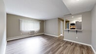 This single-floor unit is designed with a functional, open-concept layout that connects the living and dining areas seamlessly. The main living space features durable wood-look laminate flooring in a warm neutral tone, complemented by taupe-painted walls and crisp white baseboards and trim. A large window fitted with vertical blinds allows natural light to fill the room, while a wall-mounted air conditioning unit and baseboard heating system provide year-round climate control.

The kitchen is accessed via a half-wall with a breakfast bar overhang, creating an efficient pass-through that maintains visual connectivity with the living area. It is equipped with stainless steel appliances, including a gas range, over-the-range microwave, and refrigerator. The cabinetry features a two-tone design, with white upper cabinets and slate-blue lower cabinets, paired with light countertops and a double-basin stainless steel sink.

The bedrooms are carpeted in a neutral beige tone and feature vertical-blind windows that bring in natural light and outdoor greenery views. Baseboard heating runs along the perimeter of each room. The overall aesthetic throughout the unit is clean, contemporary, and move-in ready.