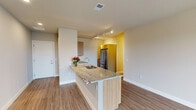 This contemporary single-floor residence offers a thoughtfully designed open-concept layout with a clean, modern aesthetic throughout. The kitchen serves as a central highlight, featuring a large granite-topped island with an undermount sink, warm wood-toned cabinetry, stainless steel appliances including a French door refrigerator and built-in microwave, and track lighting overhead. Wood-look flooring runs throughout the main living areas, adding warmth and continuity to the space.

The living area is bright and airy, bathed in natural light from large sliding glass doors that open onto a private balcony with views of the surrounding greenery. Track lighting provides additional illumination, complementing the neutral wall tones.

The bedrooms are carpeted for comfort, with one room featuring a ceiling fan and a wide window that allows ample natural light. The two bathrooms are finished with matching granite countertops and wood-toned vanities. One bathroom includes a glass-enclosed walk-in shower with built-in shelving, while the other offers a full soaking tub and shower combination. Both feature tile flooring and well-lit vanity mirrors.

Overall, the space presents a cohesive, contemporary design with quality finishes and a comfortable, functional flow throughout.