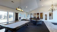 This single-floor amenity space is designed to offer both entertainment and relaxation in a well-appointed, open-concept layout. The room immediately draws attention with its vaulted ceilings, which lend an airy, expansive feel to the interior. A full-size billiards table takes center stage, positioned beneath a striking pendant light fixture adorned with candle-style bulbs that adds a warm, decorative focal point overhead. Recessed lighting throughout ensures the space is well-lit at all times.

The lounge area is furnished with plush, tufted leather sofas and armchairs arranged around a central coffee table, creating an inviting setting for socializing. A dark, patterned area rug grounds the seating arrangement and complements the room's sophisticated aesthetic. White columns and glass-paneled interior partitions divide the space into distinct zones while maintaining a sense of openness and visual connectivity between areas.

Large windows line the walls, flooding the room with natural light and offering views of the surrounding exterior. The crisp white walls and trim contrast elegantly with the darker furnishings and flooring, giving the space a clean, polished appearance. An adjoining room, visible through the glass partitions, provides additional seating and gathering space, further enhancing the versatility of this communal area.
