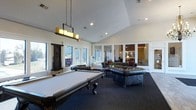 This single-floor amenity space is designed to offer both entertainment and relaxation in a well-appointed, open-concept layout. The room immediately draws attention with its vaulted ceilings, which lend an airy, expansive feel to the interior. A full-size billiards table takes center stage, positioned beneath a striking pendant light fixture adorned with candle-style bulbs that adds a warm, decorative focal point overhead. Recessed lighting throughout ensures the space is well-lit at all times.

The lounge area is furnished with plush, tufted leather sofas and armchairs arranged around a central coffee table, creating an inviting setting for socializing. A dark, patterned area rug grounds the seating arrangement and complements the room's sophisticated aesthetic. White columns and glass-paneled interior partitions divide the space into distinct zones while maintaining a sense of openness and visual connectivity between areas.

Large windows line the walls, flooding the room with natural light and offering views of the surrounding exterior. The crisp white walls and trim contrast elegantly with the darker furnishings and flooring, giving the space a clean, polished appearance. An adjoining room, visible through the glass partitions, provides additional seating and gathering space, further enhancing the versatility of this communal area.