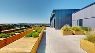 Located at 45 Gorge Rd E in Victoria, British Columbia, this rooftop terrace offers a striking outdoor amenity space defined by contemporary design and sweeping panoramic views. The terrace is clad in modern corrugated metal panels in muted blue-grey tones, lending an industrial-modern aesthetic that complements its urban setting.

The generous open-air deck is finished with large-format porcelain tiles laid in a clean grid pattern, providing a polished and durable surface throughout. A sleek black metal and glass railing system lines the perimeter, ensuring unobstructed sightlines across the Victoria skyline, surrounding greenery, and distant mountain ranges beyond.

A standout feature of the terrace is its series of raised wooden planter boxes, constructed from warm-toned timber and filled with lush herbs, grasses, and ornamental plantings. These garden beds introduce a natural, living element that softens the industrial architecture and creates a welcoming, park-like atmosphere.

The space is organized to accommodate multiple uses, with areas suited for casual seating and social gatherings, complemented by tall bistro-style tables near the building entrance. A partially covered pergola section provides shade, enhancing year-round usability. Bathed in abundant natural light and open to clear skies, this rooftop terrace delivers an exceptional outdoor experience above the Gorge neighbourhood.