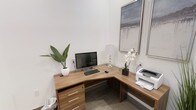 This single-floor space is designed as a functional and stylish private workspace. The room centers around a warm walnut-finish L-shaped desk that provides ample surface area for a desktop computer, printer, and personal accessories, making it well-suited for both administrative and creative tasks. The desk includes built-in drawers with sleek metal hardware, offering practical storage solutions without compromising the clean aesthetic of the space.

The walls are painted in crisp white, contributing to a bright and open atmosphere. Two framed abstract artworks are displayed on one wall, adding a polished, professional touch to the environment. Decorative elements — including a potted tropical plant and a white orchid arrangement — introduce natural warmth and visual interest to the workspace.

The flooring features neutral-toned tile, which complements the overall modern design. A crystal-based table lamp provides supplemental lighting, enhancing the room's functionality during varied lighting conditions. The space is efficiently laid out, making thoughtful use of its footprint while maintaining an uncluttered, organized feel throughout.
