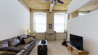 This single-floor space showcases a thoughtfully designed living area that combines rustic charm with modern comfort. The standout architectural feature is the warm, natural wood-planked ceiling, which adds texture and character to the room while housing a ceiling fan for year-round comfort. Elevated ceiling height enhances the sense of openness and volume throughout the space.

Two large arched windows dominate the far wall, flooding the room with abundant natural light and contributing to the airy atmosphere. Roman shades offer privacy while maintaining the clean, classic aesthetic. The neutral beige walls and light carpeted flooring create a soft, cohesive backdrop that complements the room's furnishings.

The living area is anchored by a rich leather sofa accented with decorative pillows, paired with a vintage-style steamer trunk serving as a coffee table—adding an eclectic, well-traveled character to the space. A wrought-iron console table beneath the windows provides functional elegance, while a flat-screen television and rustic media cabinet complete the entertainment setup. Overall, the space exudes a warm, welcoming ambiance with a tasteful mix of industrial and traditional design sensibilities.