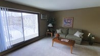 This single-floor space presents a well-appointed living area characterized by warm, earthy olive-toned walls that create a cozy, grounded atmosphere. The room is anchored by plush wall-to-wall carpeting in a neutral light gray, adding softness underfoot and complementing the overall color palette.

A large sliding glass door serves as a prominent architectural feature, flooding the space with natural light and providing direct access to an outdoor balcony. The view beyond reveals a winter streetscape with bare trees, reinforcing a sense of connection to the outdoors. A wall-mounted air conditioning unit sits adjacent to the sliding door, offering climate control convenience.

The living area is comfortably furnished with a sage green upholstered sofa accented by decorative pillows, paired with a classic wooden coffee table. A side table with a traditional lamp adds warm ambient lighting, while a lush potted plant in the corner brings a natural element to the interior. Framed artwork on the wall contributes a refined finishing touch. A baseboard heating unit runs along the exterior wall, ensuring year-round comfort in this inviting, functional space.
