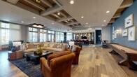 This single-floor clubhouse at Parc at Murfreesboro is a thoughtfully designed community amenity space that blends upscale aesthetics with a warm, welcoming atmosphere. The entry foyer makes an immediate impression with its striking geometric-patterned hardwood flooring, soaring ceilings, and floor-to-ceiling glass walls that bathe the space in abundant natural light while framing views of the outdoor pool and grounds.

The main lounge area features a sophisticated mix of coffered ceilings with exposed wood beams, recessed lighting, and an eclectic yet cohesive furniture arrangement of plush sofas, leather armchairs, and area rugs. Bold teal accent walls, curated artwork, and decorative pendant lighting add layers of visual interest throughout.

A full catering kitchen anchors one wing of the space, outfitted with an expansive quartz-topped island, dark cabinetry with tile detailing, and globe pendant lights. Adjacent to the kitchen, a dedicated game room offers a billiards table, bar-height seating, and built-in shelving, while a separate card room with a round game table, teal upholstered chairs, and dramatic dark walls provides an intimate setting. High-top communal tables positioned along large windows offer additional flexible seating. The seamless flow between zones, consistent hardwood flooring, and abundant glazing create a cohesive, light-filled environment throughout.