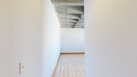 Located at 1300 Convention Plaza in downtown St. Louis, this urban loft showcases a distinctive industrial aesthetic that blends raw architectural character with functional living spaces. The open-concept main living area immediately impresses with its exposed concrete ceiling, visible steel beams, and large ductwork — hallmarks of classic loft design. Light hardwood-style flooring runs throughout the main areas, complemented by ceiling fans and track lighting that add both comfort and practicality. A bank of oversized windows floods the living space with natural light while offering views of the surrounding cityscape.

The kitchen features warm honey-toned cabinetry, granite-style countertops, and a full suite of stainless steel appliances, including a refrigerator, electric range, and dishwasher. Adjacent to the main living area, the bedroom continues the industrial aesthetic with exposed ceiling elements and generous windows fitted with blinds, providing privacy while preserving natural light. Soft carpeting in the bedroom offers a comfortable contrast to the hard flooring found elsewhere.

The bathroom is cleanly appointed with a full tub-shower combination, a single vanity with a wide mirror, and tile flooring. Additional rooms provide flexible, open spaces ideal for a home office or den, each maintaining the same exposed-ceiling character found throughout this architecturally compelling urban residence.