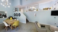 This thoughtfully designed single-floor commercial space offers a warm and professional atmosphere, characterized by its distinctive split-level layout. The interior is finished with soft, calming blue-gray walls that lend an inviting and approachable feel throughout. A staircase connects the lower level to an elevated mezzanine-style area, which is open to the main floor below through a series of white-framed partition windows, allowing natural light and sightlines to flow freely between levels. The upper area features bright overhead lighting and is tastefully decorated with small plants and seasonal accents, creating a pleasant, lived-in quality. The main level serves as a welcoming reception or waiting area, furnished with a mix of modern seating including a gray upholstered chair with a patterned pillow and vibrant yellow accent chairs alongside a wooden table. Framed artwork and circular wall mirrors add decorative character to the walls. The flooring throughout is a neutral, patterned carpet tile that complements the overall color palette. High ceilings on the lower level enhance the sense of openness, while recessed lighting ensures the space remains bright and functional. Overall, this space balances professionalism with a warm, approachable ambiance.