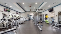 This fitness center offers a comprehensive workout environment designed to accommodate a wide range of exercise routines. The space features a tray ceiling with navy blue trim accents, recessed lighting, and red ceiling fans that contribute to both the aesthetic appeal and ventilation of the room. Durable rubber flooring covers the entire area, providing a safe and practical surface suited for all types of physical activity.

The cardio zone is well-stocked with multiple treadmills and elliptical machines, positioned along a wall of large windows that allow natural light to fill the room and offer views of the surrounding outdoor greenery. Wall-mounted flat-screen televisions are positioned throughout the space for entertainment during workouts.

The strength training area features a multi-station cable machine, a functional trainer, weight benches, and a full rack of dumbbells in varying sizes. A large wall mirror enhances the sense of depth and allows users to monitor their form. Functional fitness accessories, including stability balls and medicine balls, are neatly stored along the walls.

The space is bright, open, and thoughtfully organized to support both cardio and strength-focused training. Natural light, combined with recessed overhead lighting, ensures excellent visibility throughout the facility at all times.