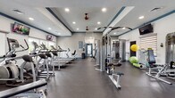 This fitness center offers a comprehensive workout environment designed to accommodate a wide range of exercise routines. The space features a tray ceiling with navy blue trim accents, recessed lighting, and red ceiling fans that contribute to both the aesthetic appeal and ventilation of the room. Durable rubber flooring covers the entire area, providing a safe and practical surface suited for all types of physical activity.

The cardio zone is well-stocked with multiple treadmills and elliptical machines, positioned along a wall of large windows that allow natural light to fill the room and offer views of the surrounding outdoor greenery. Wall-mounted flat-screen televisions are positioned throughout the space for entertainment during workouts.

The strength training area features a multi-station cable machine, a functional trainer, weight benches, and a full rack of dumbbells in varying sizes. A large wall mirror enhances the sense of depth and allows users to monitor their form. Functional fitness accessories, including stability balls and medicine balls, are neatly stored along the walls.

The space is bright, open, and thoughtfully organized to support both cardio and strength-focused training. Natural light, combined with recessed overhead lighting, ensures excellent visibility throughout the facility at all times.